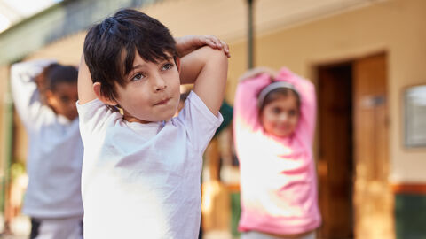 Dehnübungen zur Gymnastik Kinder dehnen ihre Arme im Sport. Ganz vorne steht ein kleiner Junge mit sehr konzentriertem Gesichtsausdruck.
