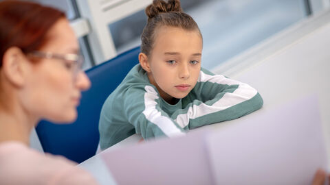Sorgen und Ängste mit einer Vertrauensperson teilen. Ein Kind sitzt betrübt an einem Tisch. Eine erwachsene Person sitzt gegenüber und zeigt ihm ein großes Blatt Papier.