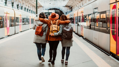 Drei Freunde sieht man von hinten wie sie Arm in Arm neben den Zügen am Bahngleis entlang laufen.