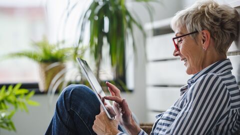 Eine ältere Dame sitzt auf dem Boden, die Beine angewinkelt und ein Tablet in der Hand.