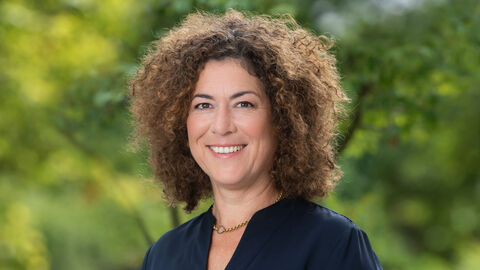 Alice Engel Alice Engel lächelt in die Kamera. Sie trägt eine goldene Kette und eine dunkelblaue Bluse. Im Hintergrund sind grüne Pflanzen zu sehen.