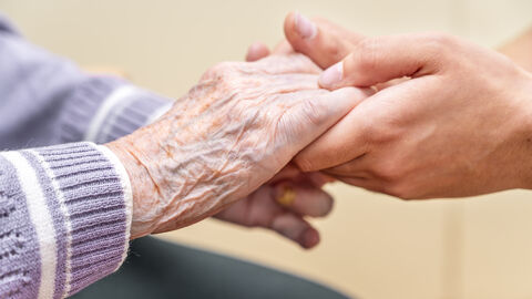 Es sind die Hände von zwei Menschen zu sehen. Die linken Hände (von einer Seniorin oder Senior) legt seine/ihre Hände in die einer jüngeren Person