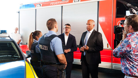 Ministerpräsident Rhein und Innenminister Poseck im Dialog mit Einsatzkräften Ministerpräsident Rhein und Innenminister Poseck stehen vor einem Feuerwehrfahrzeug und unterhalten sich mit zwei Polizisten.