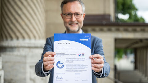 Museumsdirektor des Landesmuseums Wiesbaden, Dr. Andreas Henning, hält das Zertifikat als klimaneutrale Organisation in die Kamera.
