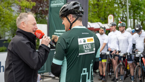 Ministerpräsident Rhein in seinem Raddress neben einem Mann mit Mikrofon, beide unterhalten sich