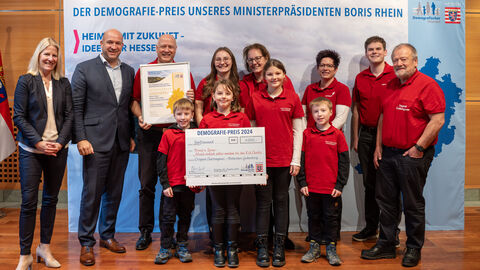 Eine Gruppe Personen steht vor einem Aufsteller mit der Aufschrift "Der Demografie-Preis unseres Ministerpräsidenten Boris Rhein". Eine Person hält eine gerahmte Urkunde in Händen, eine weitere einen Scheck.